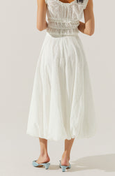A woman is wearing a white eyelet dress with a sleeveless top featuring ruffled details and a gathered waist. The skirt is full-length, flowing, and also made of eyelet fabric, creating a soft, airy appearance. She is wearing light blue open-toe heels with a mid-height heel.